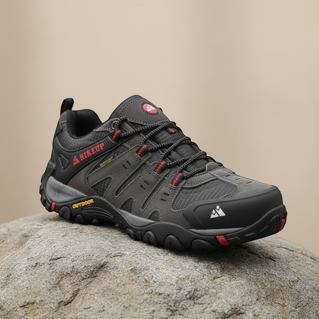Vue de profil d'une chaussure de randonnée technique pour homme, associant des empiècements en daim gris anthracite et en textile respirant, avec des accents de lacets rouges et une semelle à crampons, reposant sur un rocher.